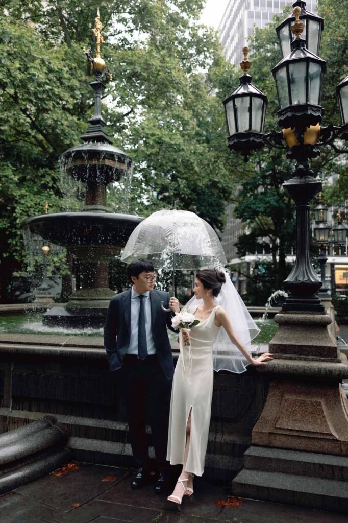 Bride and groom sharing a romantic moment under a clear umbrella by a fountain near New York City Hall on a rainy day