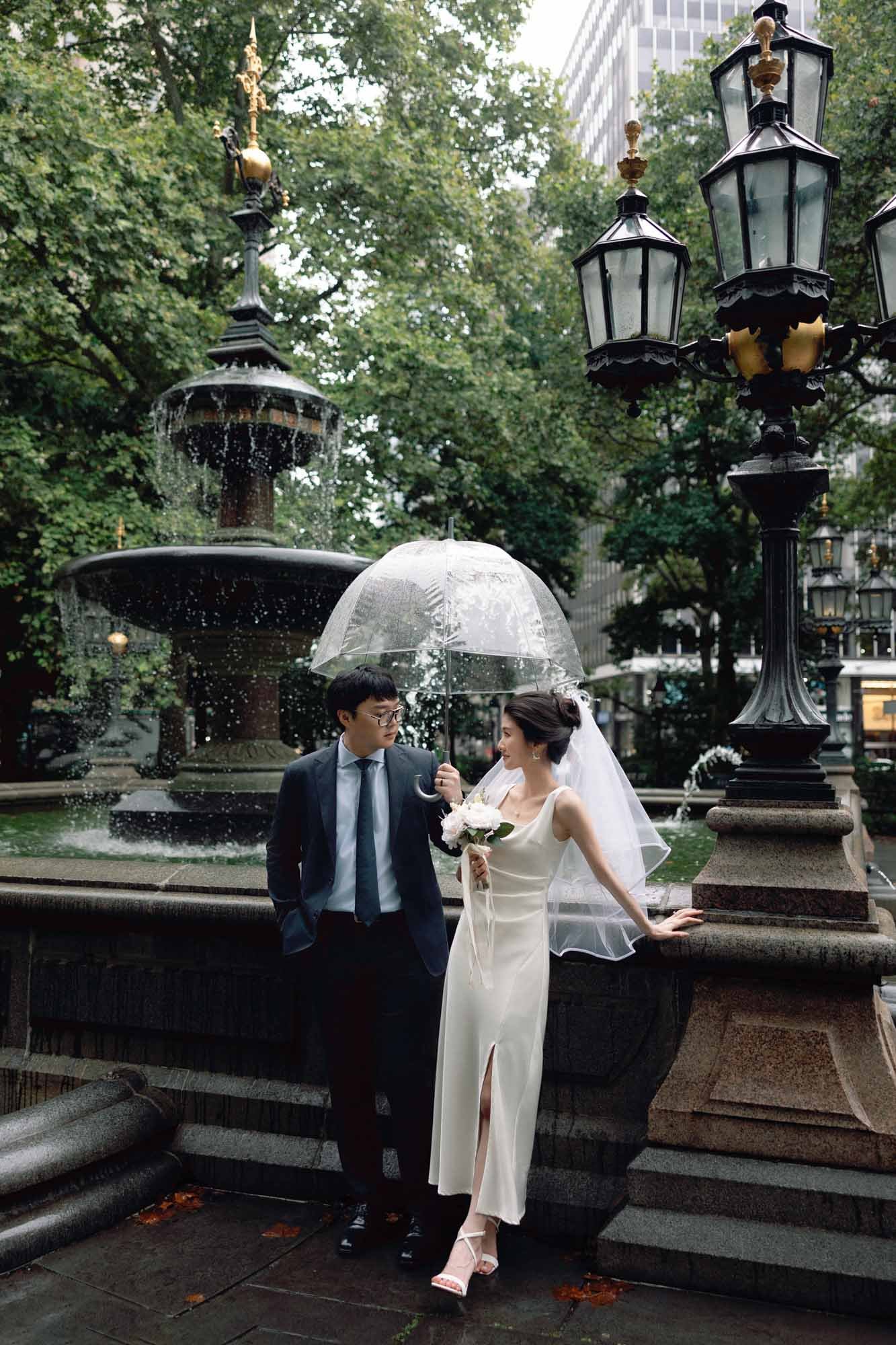 Bride and groom sharing a romantic moment under a clear umbrella by a fountain near New York City Hall on a rainy day