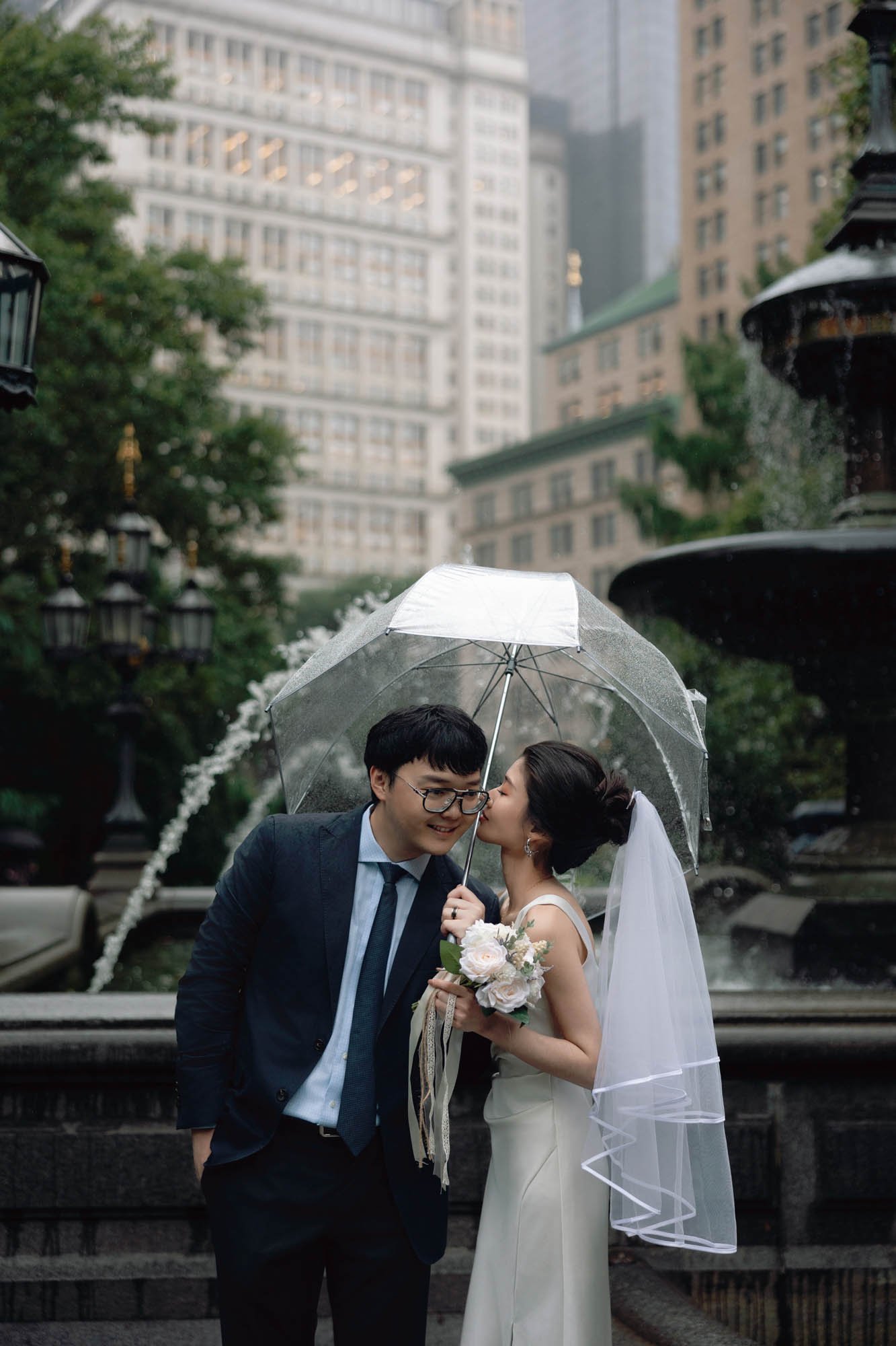 Bride and groom sharing a romantic moment under a clear umbrella by a fountain near New York City Hall on a rainy day